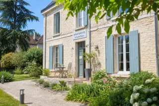 Charming guesthouse facade with sunny stone walls, lush summer garden, and inviting bistro table under flowering pergola